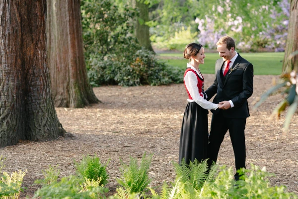 The bride in a Norwegian bunad and the groom in a dark suit stand facing each other on a garden path, smiling and holding hands surrounded by ferns and trees. Candid documentary wedding portrait.