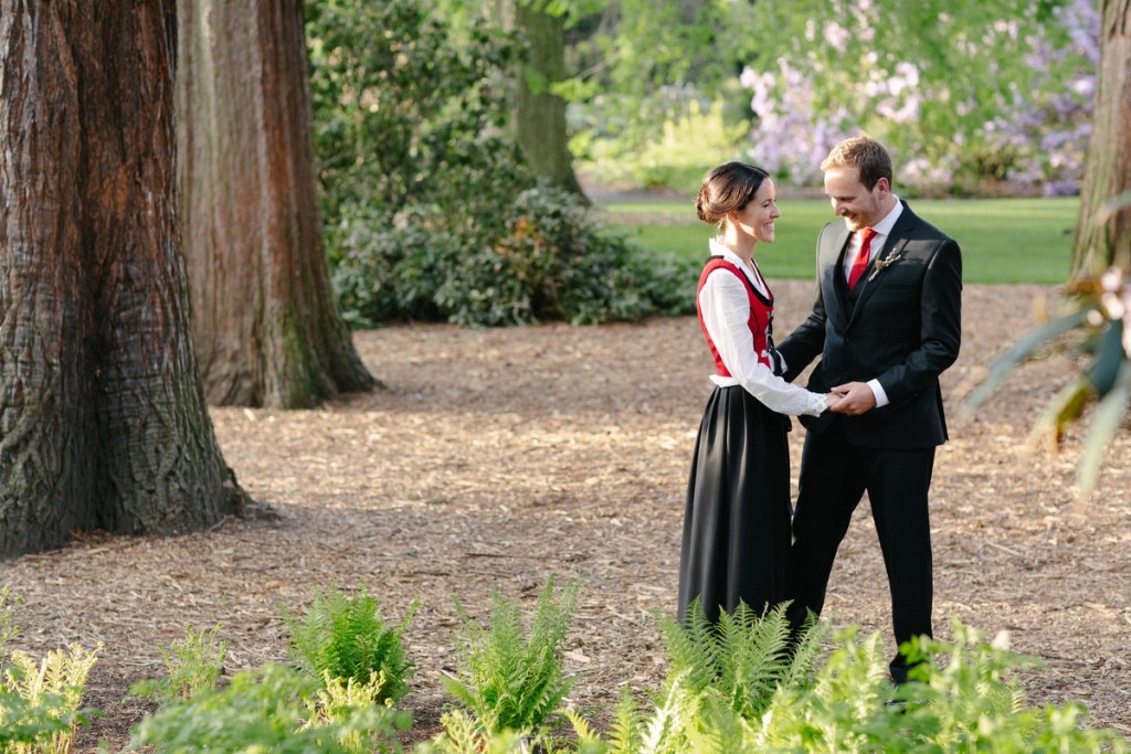 The bride in a Norwegian bunad and the groom in a dark suit stand facing each other on a garden path, smiling and holding hands surrounded by ferns and trees. Candid documentary wedding portrait.