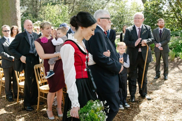 Bride in a red Norwegian bunad walking down the outdoor aisle arm in arm with her father, guests watching from either side among tall trees. Candid wedding processional.