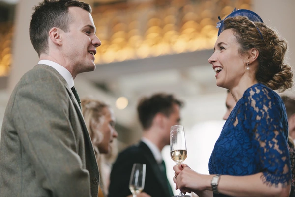 Man and woman chatting cheerfully at the wedding reception — blue lace dress, grey suit, white wine