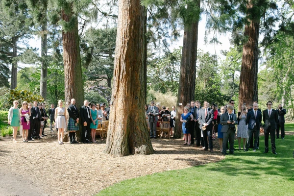 Wide candid shot of wedding guests gathered among giant redwood trees at the Royal Botanic Garden Edinburgh, awaiting the ceremony. Documentary wedding photography.