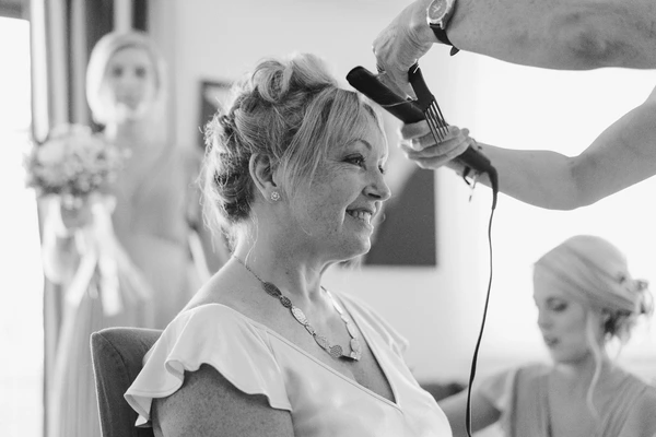 Middle-aged woman smiles as a hairdresser styles her elegant wedding updo in a bright room