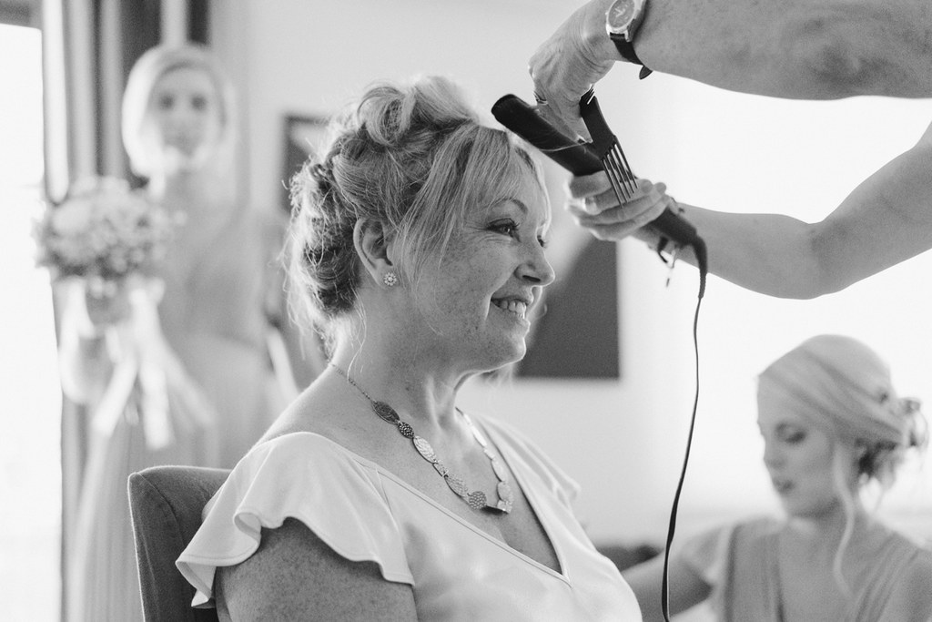 Middle-aged woman smiles as a hairdresser styles her elegant wedding updo in a bright room