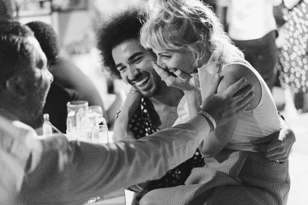 Young couple laughing together at the wedding table surrounded by guests — genuine joy. Black-and-white.