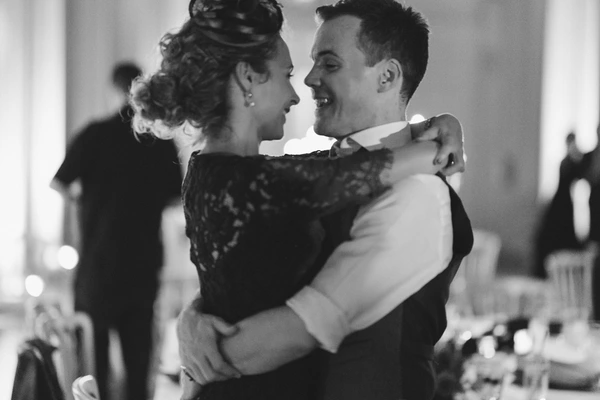 Groom lifts the bride during a wedding dance, both smiling — black lace dress, blurred guests