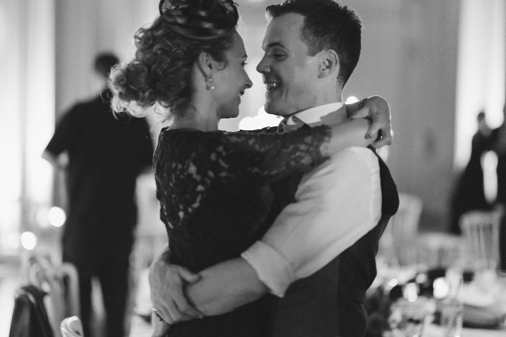 Groom lifts the bride during a wedding dance, both smiling — black lace dress, blurred guests