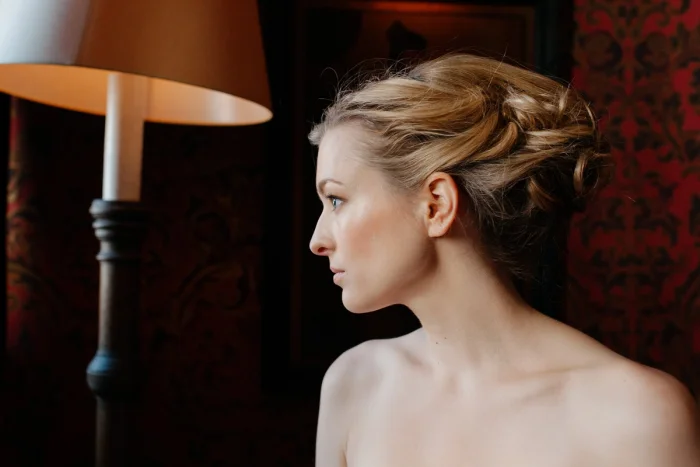 Bride in elegant updo gazes pensively in profile, warm lamplight against rich red damask wallpaper