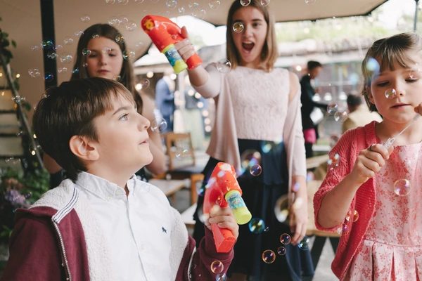 Children blow soap bubbles at the outdoor wedding — boy looking up in wonder, girl in pink dress