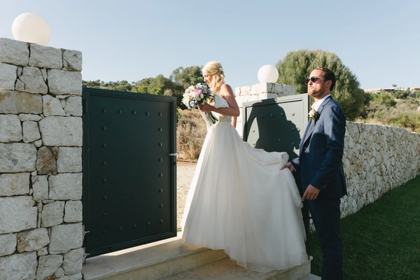 Bride and groom before a black gate with stone wall — bride holds pink bouquet, Mediterranean greenery