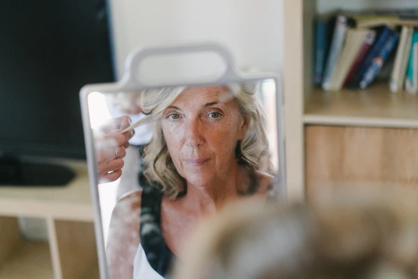 Middle-aged woman smiles while applying makeup with a brush in a mirror — home bathroom, natural light