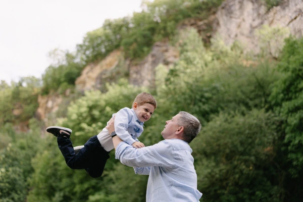 Father playfully holding son in air during wedding reception with green mountain landscape backdrop.