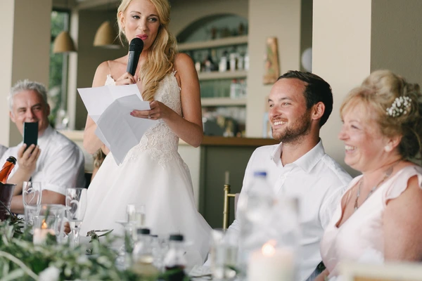 Bride with microphone at the table in white lace, groom beside her with a happy smile, guests around