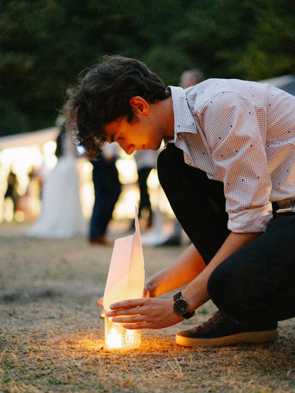 Guest lighting candle lantern at outdoor evening wedding reception with warm glow.
