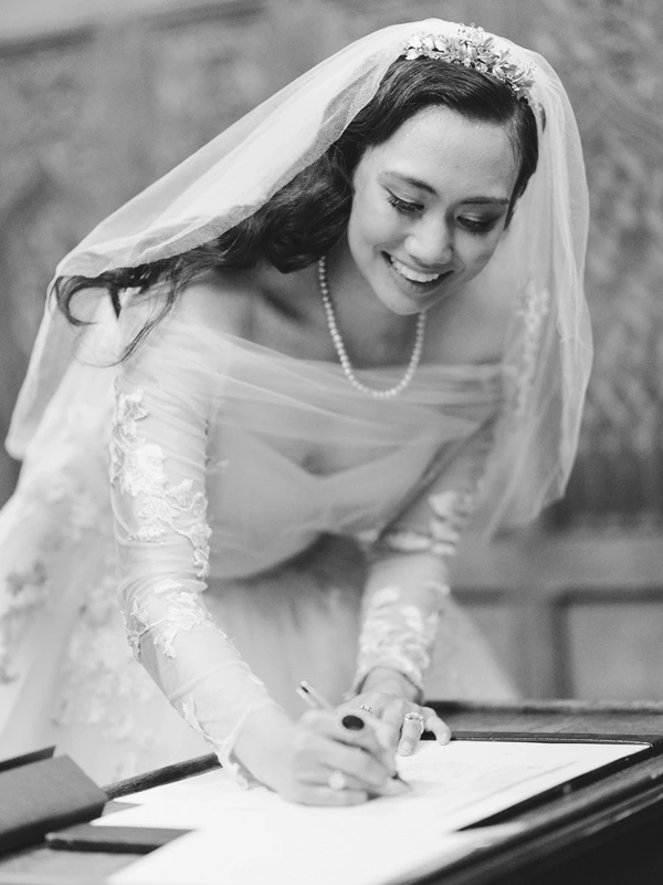 Smiling bride in lace and veil signs the register — pearl necklace and floral crown. Black-and-white.