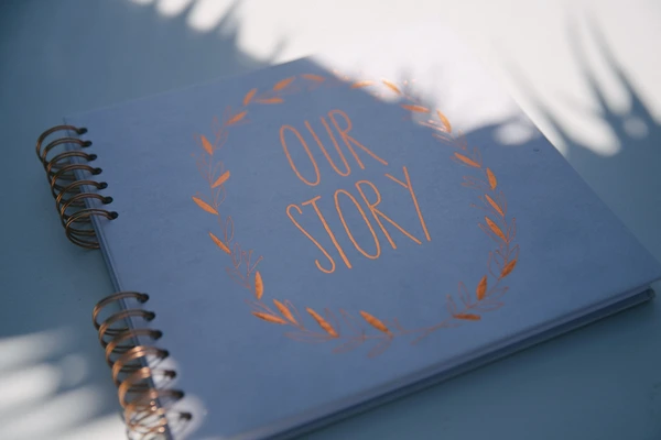 Open spiral notebook reading "Our Story" in bronze lettering with leaf wreath, sunny shadows