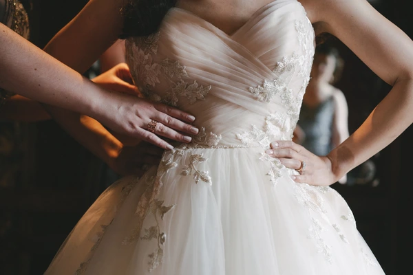Bride's white silk pearl-trimmed gown being arranged by helping hands — golden light plays on the fabric