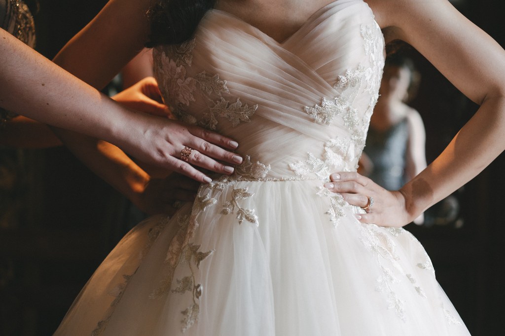 Bride's white silk pearl-trimmed gown being arranged by helping hands — golden light plays on the fabric