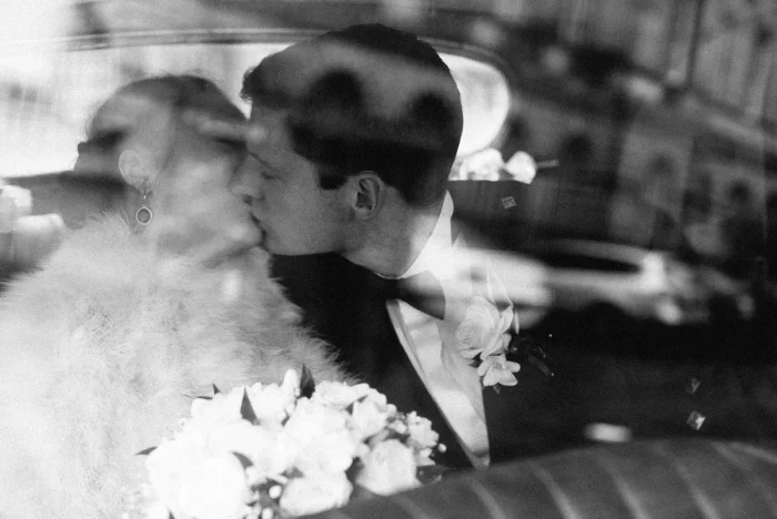 Black and white photo of newlyweds kissing in a car, bride holding bouquet, groom in tuxedo, romantic vintage mood.