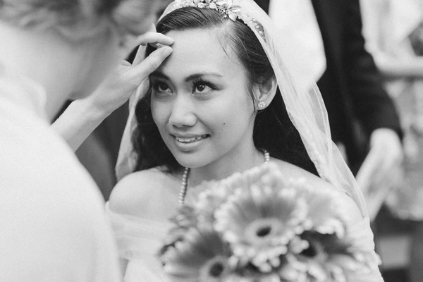 Smiling bride with white veil and pearl headband raises her hand to her brow — black-and-white