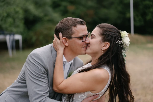 Groom kissing bride on cheek with tender moment at outdoor garden wedding under green trees.