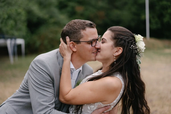Groom kissing bride on cheek with tender moment at outdoor garden wedding under green trees.