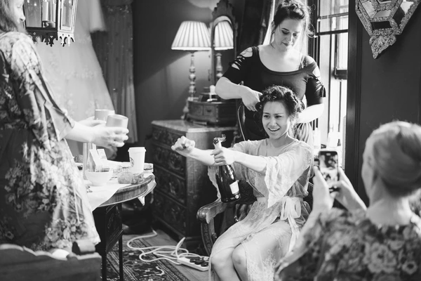 Bride in white lace sits happily holding champagne, hairdresser finishing her updo, bridesmaids and friends around her in warm natural light.