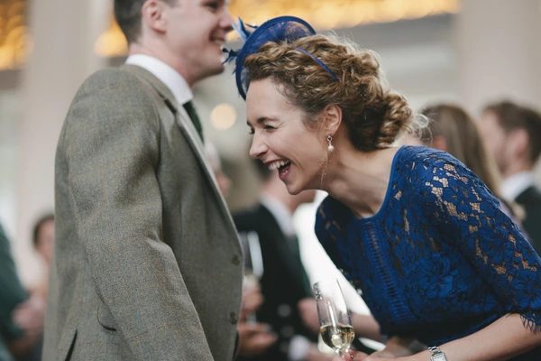 Woman in blue lace and headpiece laughing with a man in grey suit, white wine in hand