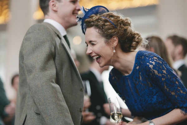 Woman in blue lace and headpiece laughing with a man in grey suit, white wine in hand