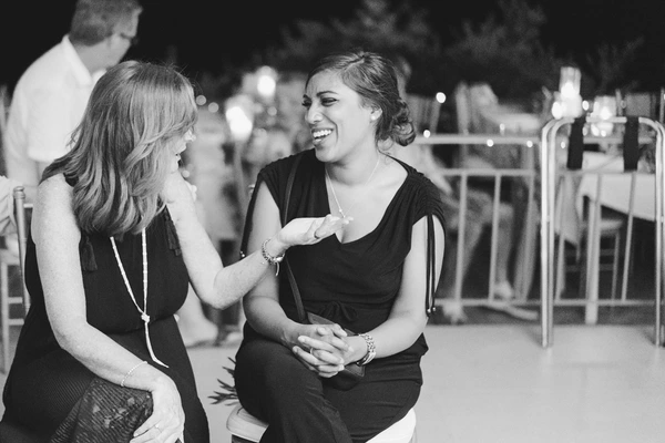 Two women chat and laugh at the outdoor evening event — black elegant dresses, intimate friend moment