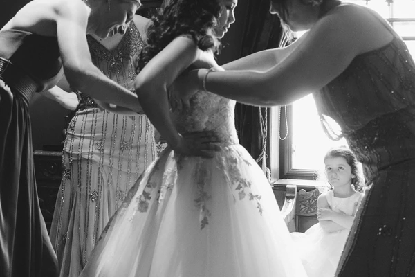 Bride stands in white lace as helpers and a little girl surround her — natural window light