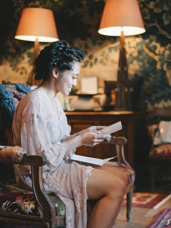 Bride in white lace reads papers in a decorated armchair before the wedding — warm lamp light