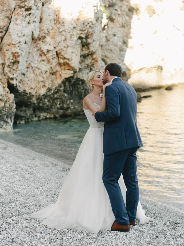Bride and groom kiss on the pebble beach beside a rocky cliff — white lace, dark navy, soft light