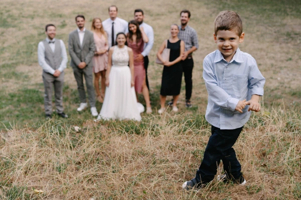 Young boy running in field with bride and wedding guests in background at outdoor celebration.