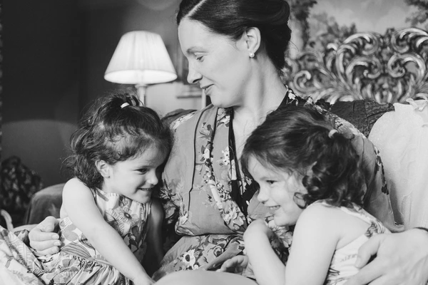 Mother and two little girls in a tender moment — smiling at each other on a sofa, warm lamp light