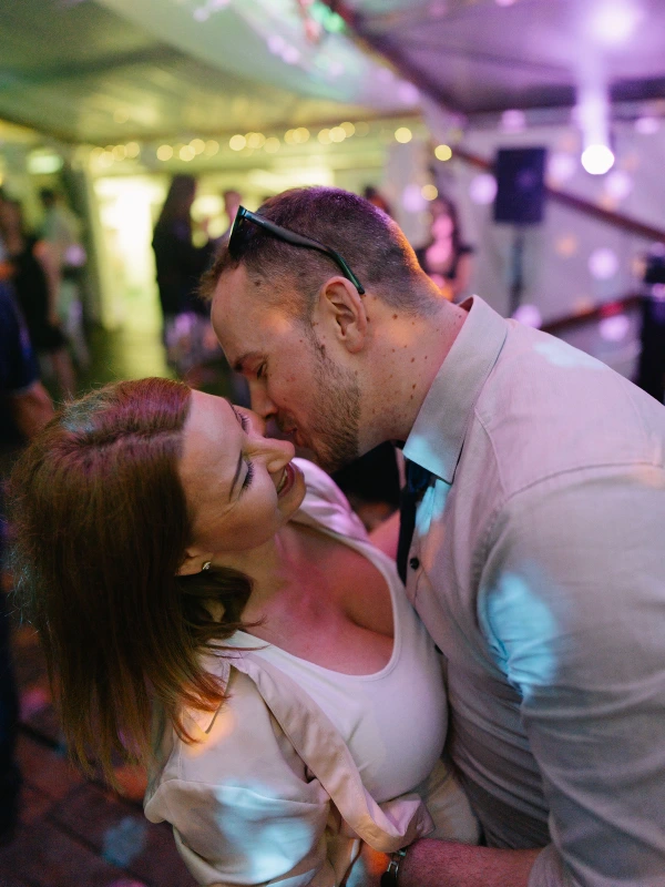 Groom and bride embracing at reception — colorful ambient lighting, intimate dancing moment, festive party atmosphere.