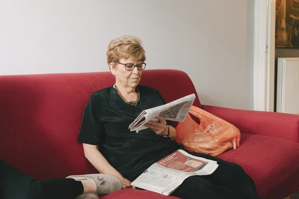 Older woman in glasses reads a newspaper on a burgundy sofa in a white-walled room