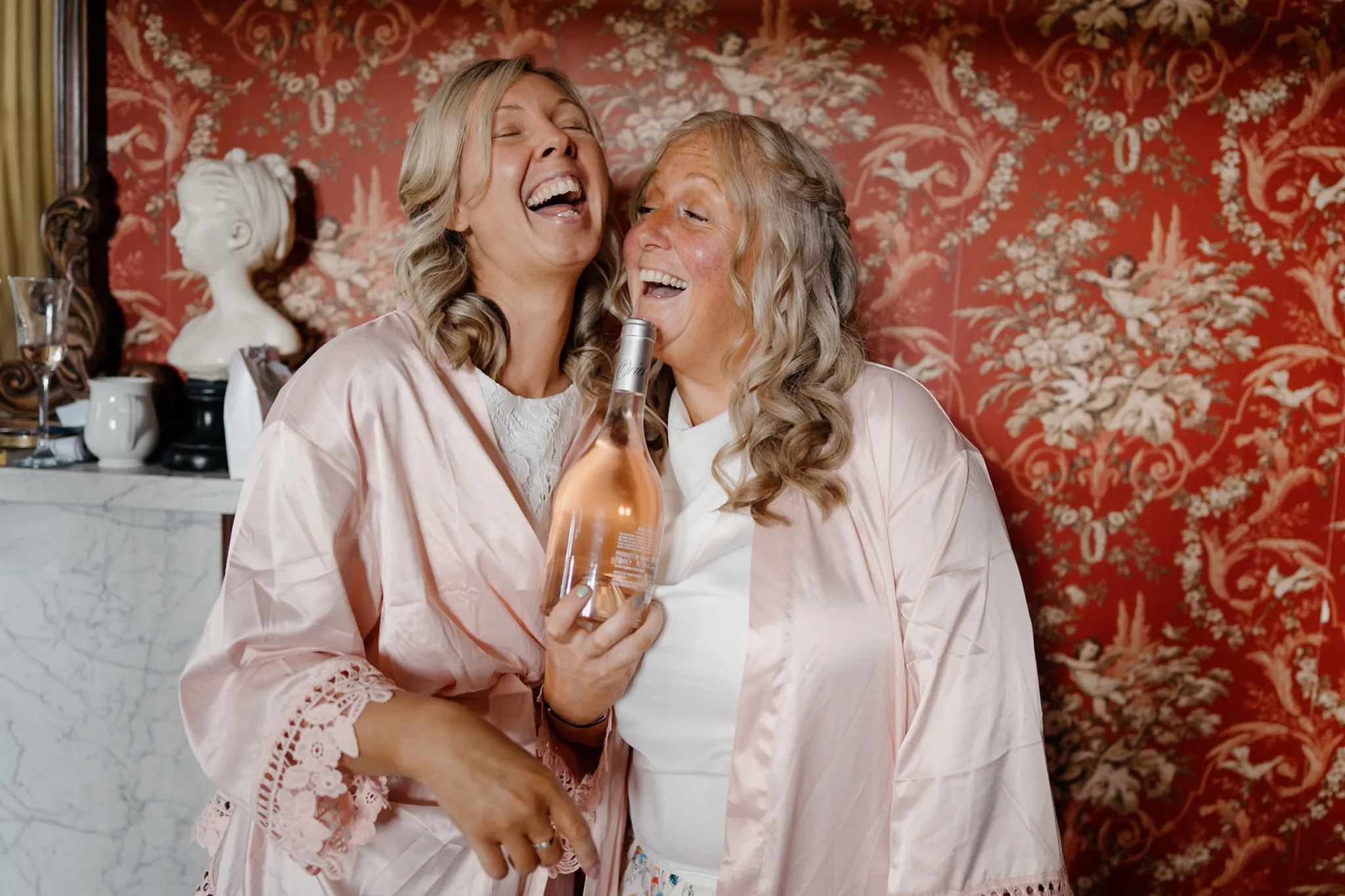 Two laughing women in pink satin robes share a bottle of rosé wine while getting ready before a wedding ceremony.