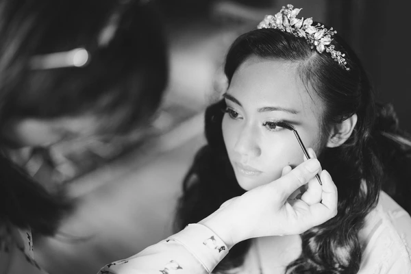 Bride with jewelled hair detail concentrates while applying makeup — black-and-white portrait