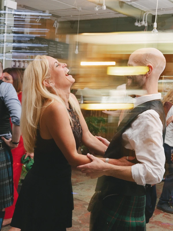 Joyful couple dances at the wedding party — woman in black, man in tartan — smiling guests
