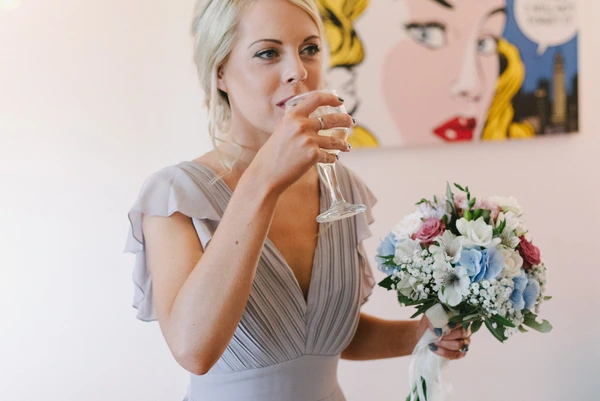 Blonde woman in grey dress holds a bouquet of white, blue and pink flowers, thoughtful expression