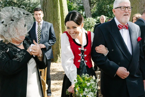 Bride in her Norwegian bunad arriving on her father's arm and greeted warmly by guests at the outdoor ceremony in the Royal Botanic Garden. Candid processional moment.