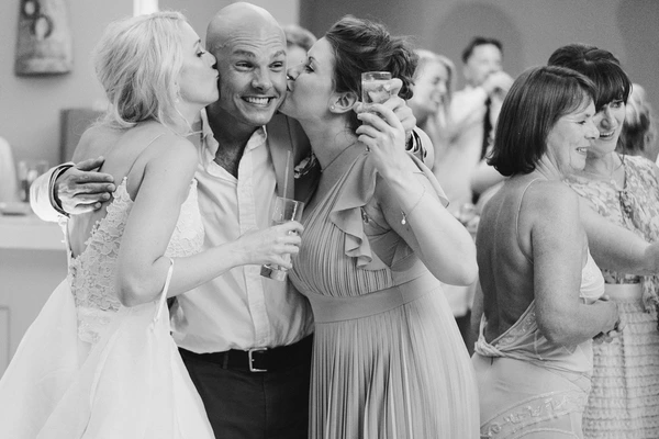 Groom laughs as two women kiss him on the cheeks at the same time — guest holds a glass, lively reception