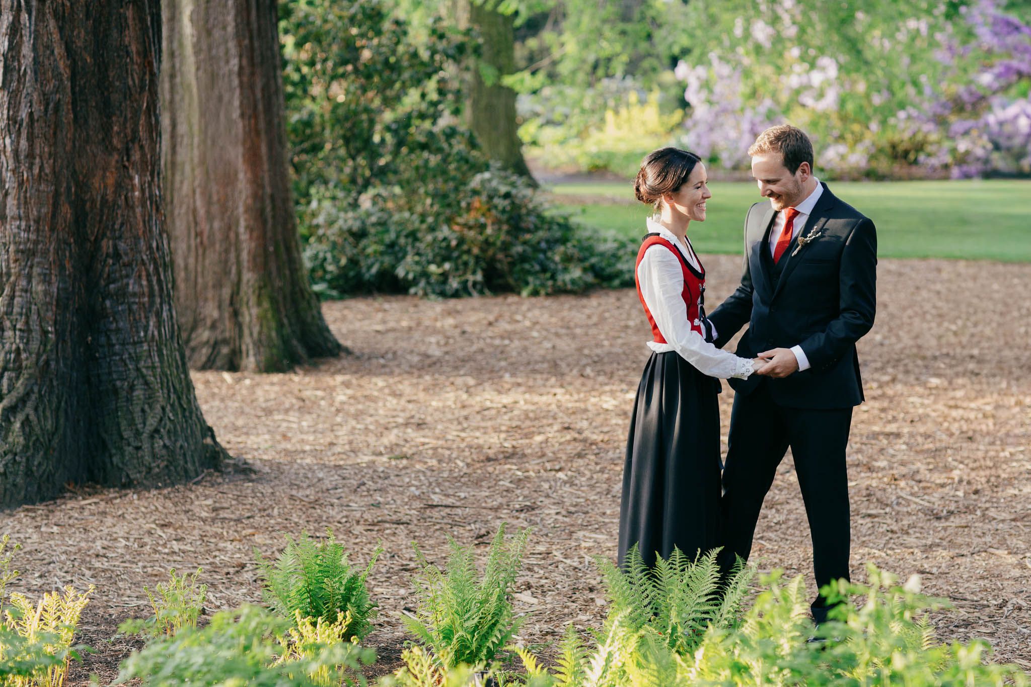 Smiling couple in formal attire holding hands in a lush Edinburgh Botanic Garden park, bride in traditional Norwegian bunad, groom in dark suit.