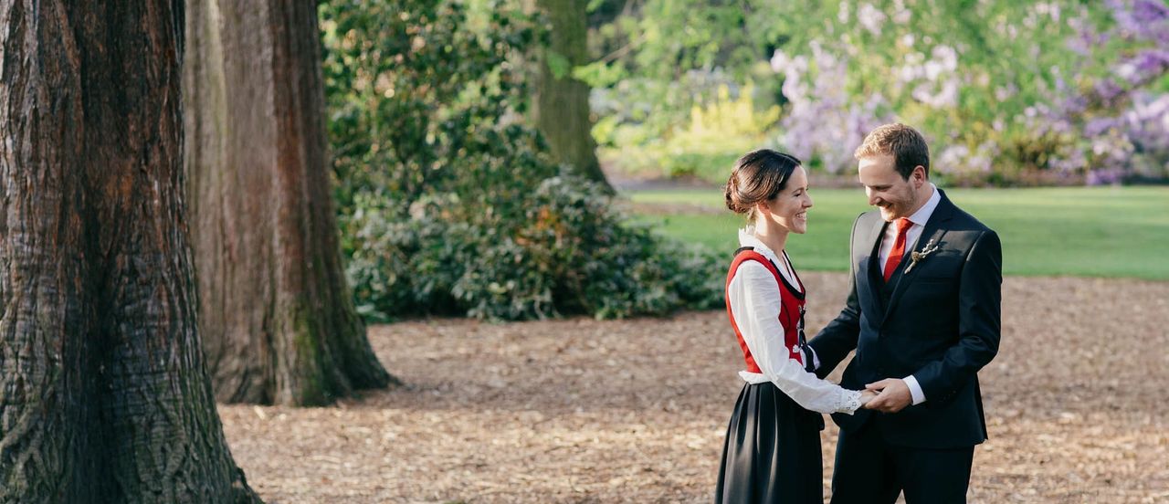 Smiling couple in formal attire holding hands in a lush Edinburgh Botanic Garden park, bride in traditional Norwegian bunad, groom in dark suit.