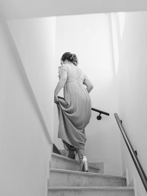 Bride walks up white stone stairs holding her ruffled skirt — bright minimal room, black-and-white