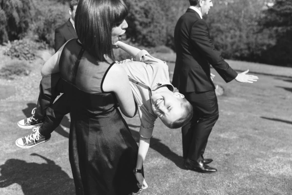 Black and white candid shot of a bridesmaid in a black dress playfully holding a laughing boy upside down in the sunlit garden, a groomsman walking past in the background. Joyful documentary moment.