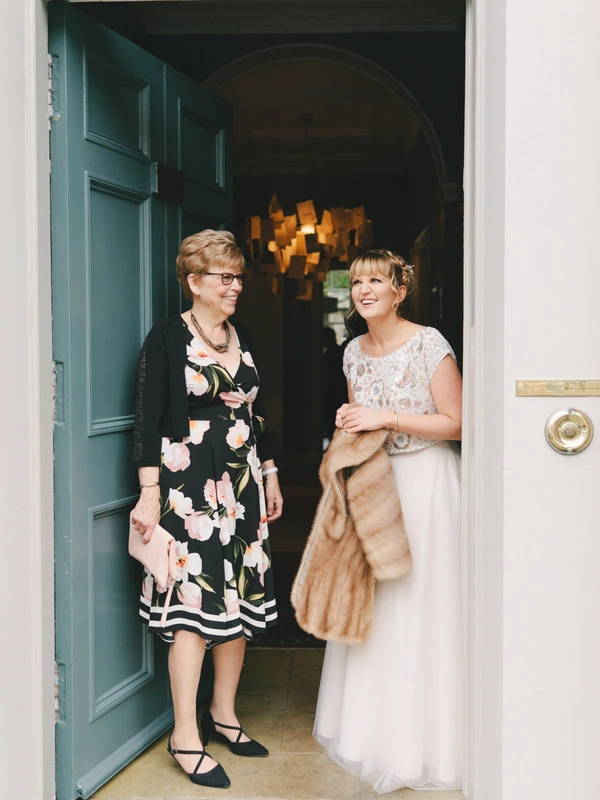 Bride in lace dress with fur stole talks to a woman in floral black dress in the doorway