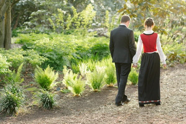 The bride in a Norwegian bunad and the groom in a dark suit walk hand in hand away from the camera through a lush garden at the Royal Botanic Garden Edinburgh. Candid documentary wedding portrait.
