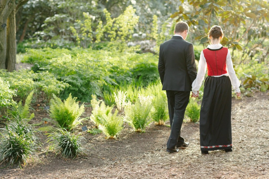 The bride in a Norwegian bunad and the groom in a dark suit walk hand in hand away from the camera through a lush garden at the Royal Botanic Garden Edinburgh. Candid documentary wedding portrait.