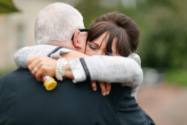 A woman with short dark hair presses her cheek against an older man's head in a close, emotional embrace at the wedding, still holding a bubble wand. Candid documentary wedding photograph.